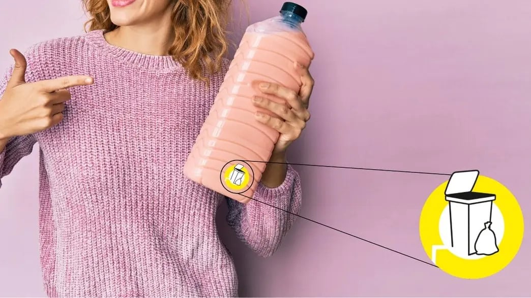 Kobieta w różowym swetrze trzyma dużą plastikową butelkę. Wskazuje palcem na piktogram recyklingowy, który jest połączony linią z ikoną kosza na śmieci, ilustrując instrukcję segregacji.