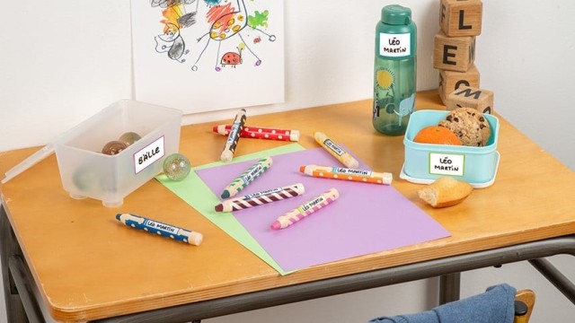 Biurko szkolne z różnymi artykułami, kostkami z literkami, rozrzuconymi kredkami oraz dwoma plastikowymi opakowaniami z naklejkami z napisem ręcznym oraz plastikową butelką z taką samą nakljeką