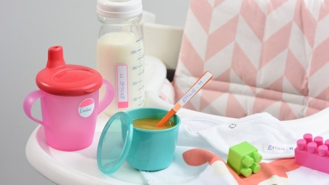Różowa butelka plastikowa z czerwonym dziubkiem, zielony pastelowy kubeczek, butelka plastikowa z mlekiem oraz różne zabawki i artykuły dziecięce na białym plastikowym stoliku 