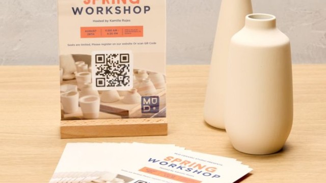 Stojąca na blacie informacja o warsztatach ceramiki „Spring Workshop” z datą, godziną i miejscem, na której centralnie umieszczono kod QR do rejestracji; obok leżą ulotki z tym samym projektem i stoją dwie jasne wazony.