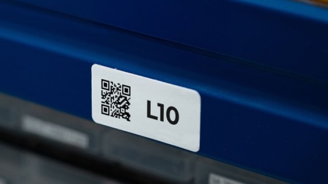 Zbliżenie na białą etykietę z kodem QR i oznaczeniem „L10”, naklejoną na niebieskim regale magazynowym.
