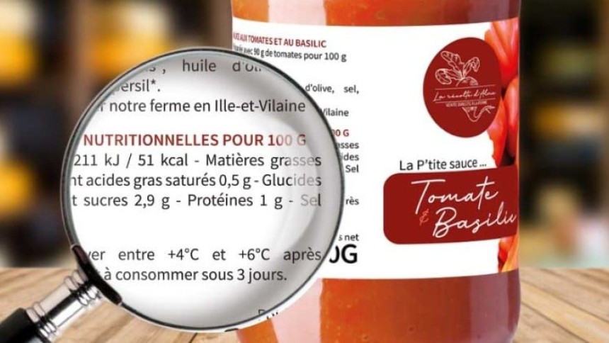  Zbliżenie etykiety sosu pomidorowego z bazylią na słoiku, tekst „Tomate & Basilic”. Przed słoikiem lupa powiększająca fragment informacji o wartościach odżywczych.