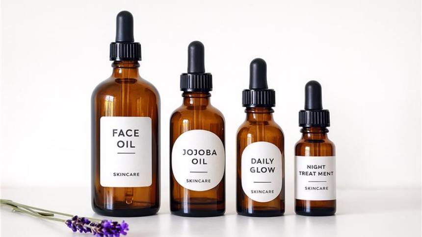 Cztery brązowe butelki z pipetą do pielęgnacji twarzy ustawione w rzędzie na białym tle; etykiety opisane kolejno: „Face Oil”, „Jojoba Oil”, „Daily Glow” i „Night Treatment”, z drobnym dopiskiem „Skincare”; z lewej leży gałązka lawendy.