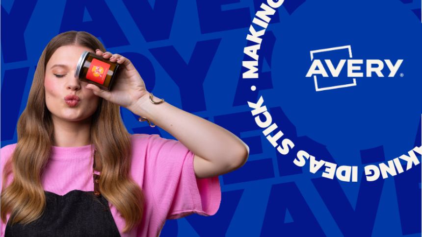 Młoda kobieta w różowej koszulce i czarnym fartuchu, z rozpuszczonymi włosami, robi "dziubek" do aparatu. W dłoni trzyma mały słoiczek z etykietą produktu, zasłaniając nim jedno oko. W tle niebieska grafika z logo i hasłem Avery: "Making Ideas Stick".