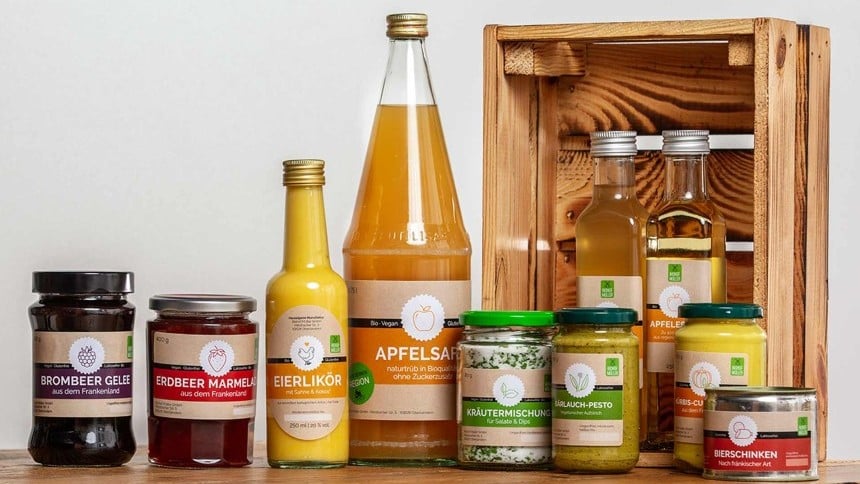 Asortyment przetworów spożywczych marki Biohof Müller. Na drewnianym stole stoją słoiki z dżemem jeżynowym i truskawkowym, pesto, mieszanką ziół, a także butelki z likierem jajecznym, sokiem jabłkowym i olejem. W tle widać drewnianą skrzynkę.