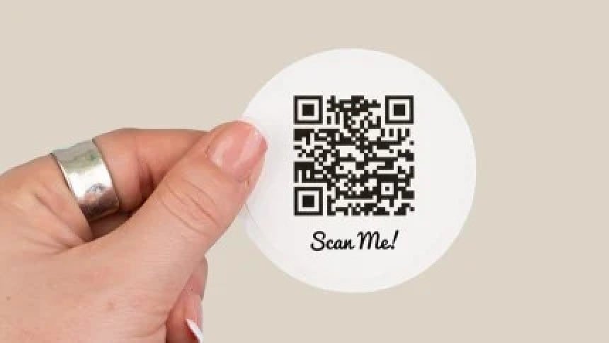 Dłoń trzymająca okrągłą białą naklejkę z kodem QR i odręcznym napisem Scan Me!