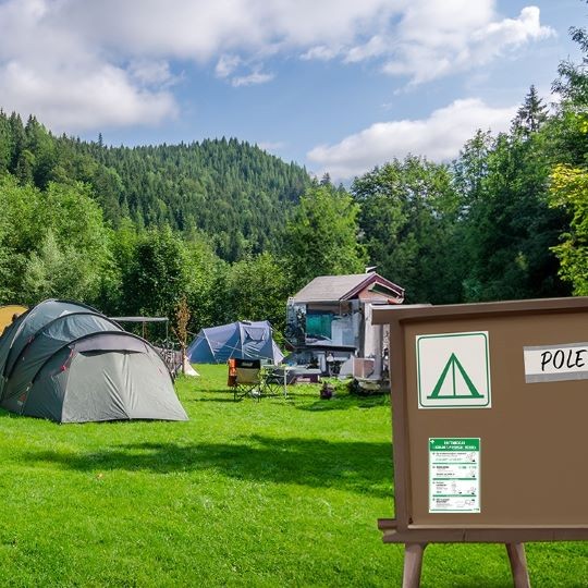Pole namiotowe z kilkoma namiotami i górami w tle, gdzie na pierwszym planie jest drewniana tablica na której są różne zielone znaki informacyjne