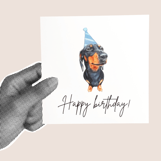 Czarnobiała dłoń trzymająca naklejkę z jamnikiem w czapce urodzinowej i napisem Happy Birthday