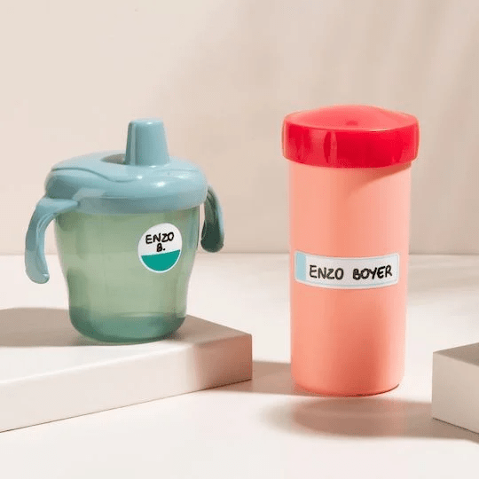 Zielony plastikowy przezroczysty kubek z dziubkiem i uchwytami oraz łososiowa podłużna butelka plastikowa z czerwoną nakrętką, na których naklejone są naklejki do opisu ręcznego