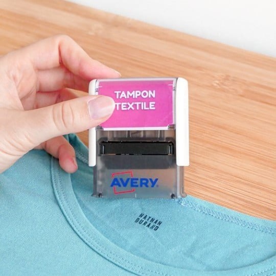 Pieczątka do odzieży Avery z napisem Tampon Textile na różowym tle naklejająca pieczątkę na kołnierzu zielonej koszulki