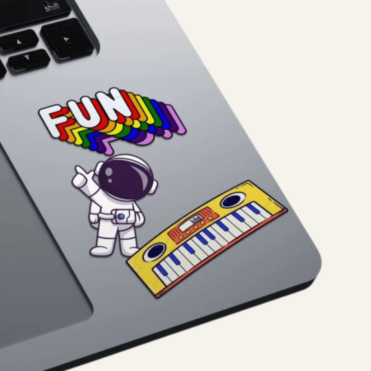 Srebrny laptop z trzema naklejkami: kolorowym napisem ‘FUN’, rysunkiem astronauty w skafandrze oraz żółtą klawiaturą muzyczną z głośnikami
