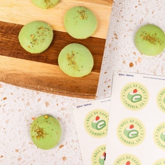 Zielone makaroniki posypane pistacjami ułożone na drewnianej desce i blacie. Obok leżą arkusze z okrągłymi etykietami z napisem ‘Roll Up Café, Cambria, CA’ oraz zieloną literą e i małą czerwoną filiżanką.