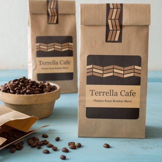 Trzy papierowe opakowania kawy marki Terrella Cafe z nadrukiem 'Medium Roast Brazilian Blend' ustawione na niebieskim tle. Obok leży otwarta torebka z kawą ziarnistą oraz miska z ziarnami kawy