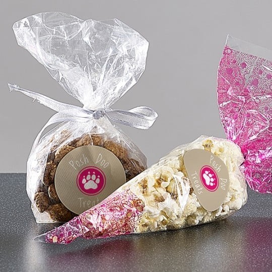 Dwa przezroczyste woreczki z psiimi smakołykami, ozdobione naklejkami z różową łapką i napisem ‘Posh Pooch Treats’. Jeden woreczek związany srebrną wstążką zawiera brązowe ciasteczka, drugi w kształcie rożka wypełniony jest popcornem