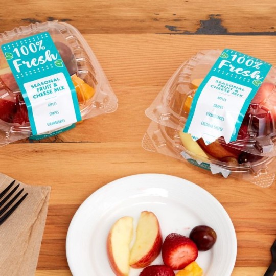 Dwa plastikowe pojemniki z plombami naklejonymi na zamknięciach, oznaczone napisami '100% Fresh' i zawierające mieszankę świeżych owoców. Na stole obok biały talerz z kawałkami jabłka, truskawką i winogronem