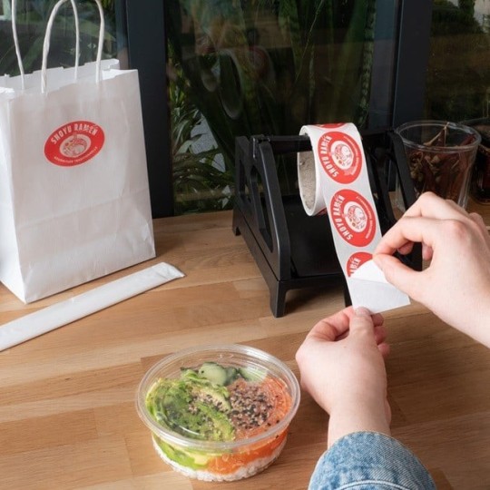 Osoba odkleja okrągłą, czerwoną naklejkę z napisem „Shoyu Ramen” z rolki na dyspenserze; obok papierowa torebka z taką samą naklejką i pojemnik z jedzeniem.