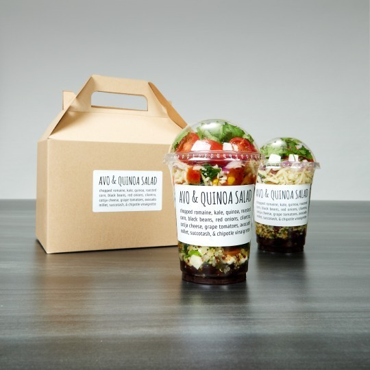 Dwie plastikowe kubki z sałatką z awokado i quinoa, każdy z etykietą „Avo & Quinoa Salad”, obok kartonowego pudełka na wynos ze zbliżoną etykietą. Zestaw ustawiony na ciemnym blacie.