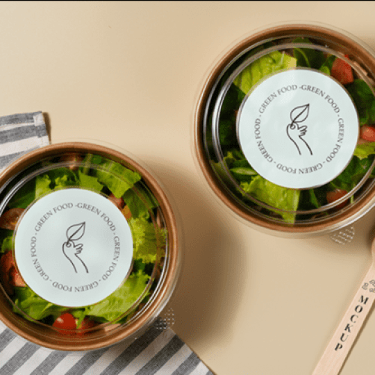 Dwa okrągłe, przezroczyste pojemniki z sałatką, z etykietą „Green Food” i rysunkiem liścia. Obok zestaw drewnianych sztućców i pasiasta serwetka, wszystko ustawione na beżowym tle.