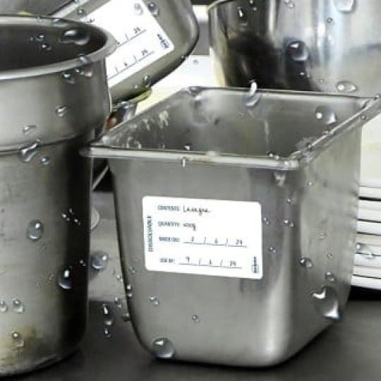 Kilka mokrych, metalowych pojemników gastronomicznych i misek na stalowym blacie, obok stosu białych talerzy. Na pojemnikach znajdują się białe etykiety. Widać krople wody, co sugeruje, że naczynia zostały niedawno umyte.