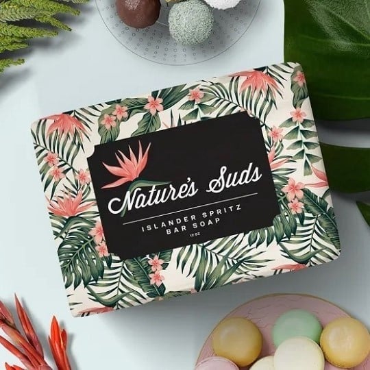 Kostka mydła owinięta papierem z kolorowym, roślinnym wzorem i etykietą „Nature’s Suds Islander Spritz Bar Soap”. W tał leżą kolorowe pralinki i liście.