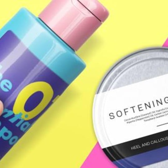 Ręka trzyma niebieską butelkę szamponu z różową zakrętką, obok okrągłe opakowanie z napisem „Softening Balm” na jasnym, żółto-różowym tle.
