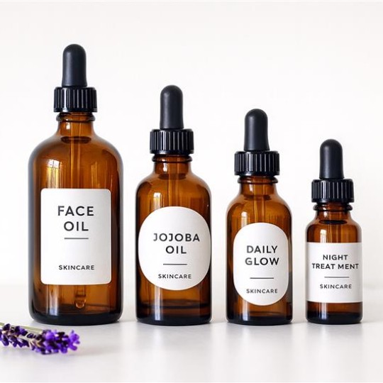 Cztery brązowe butelki z pipetą do pielęgnacji twarzy ustawione w rzędzie na białym tle; etykiety opisane kolejno: „Face Oil”, „Jojoba Oil”, „Daily Glow” i „Night Treatment”, z drobnym dopiskiem „Skincare”; z lewej leży gałązka lawendy.