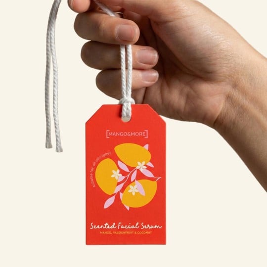 Dłoń trzyma czerwoną, prostokątną zawieszkę produktową na białym sznurku. Na zawieszce znajduje się grafika z mango i napis "Scented Facial Serum".