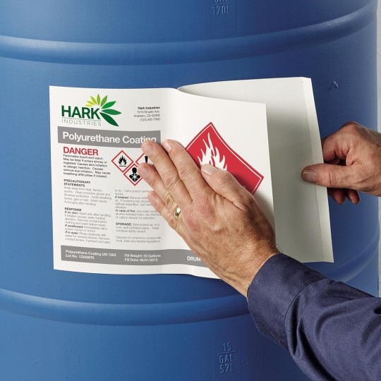 Zbliżenie na dłoń w rękawicy naklejającą dużą etykietę chemiczną GHS z piktogramem substancji łatwopalnej na niebieską, plastikową beczkę.