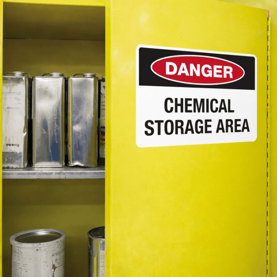 Znak bezpieczeństwa "DANGER. CHEMICAL STORAGE AREA" umieszczony na drzwiach żółtej szafy do przechowywania chemikaliów. Szafa jest częściowo otwarta, ukazując na półkach metalowe puszki i pojemniki z substancjami chemicznymi.