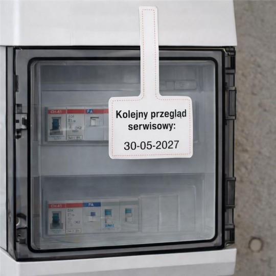 Biała zawieszka serwisowa z napisem „Kolejny przegląd serwisowy: 30-05-2027” przyczepiona do przeszklonej skrzynki z bezpiecznikami.