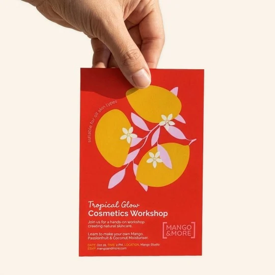 Dłoń trzymająca czerwoną ulotkę z zaproszeniem na warsztaty. Na ulotce znajduje się grafika z trzema żółtymi owocami mango i białymi kwiatami. Tekst na ulotce: "Tropical Glow Cosmetics Workshop. Dołącz do warsztatów z tworzenia naturalnych kosmetyków. Naucz się robić własny nawilżacz z mango, marakui i kokosa. Data: 26 października, godz. 14:00. Miejsce: Mango Studio. RSVP: mangoandmore.com". W rogu logo "Mango & More".