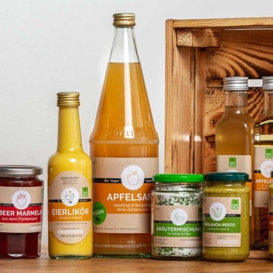 Asortyment przetworów spożywczych marki Biohof Müller. Na drewnianym stole stoją słoiki z dżemem jeżynowym i truskawkowym, pesto, mieszanką ziół, a także butelki z likierem jajecznym, sokiem jabłkowym i olejem. W tle widać drewnianą skrzynkę.