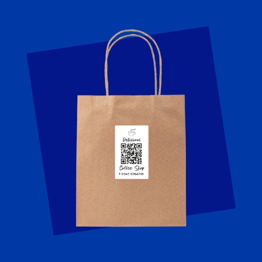 Papierowa torba na zakupy z naklejoną etykietą, na której znajduje się logo, kod QR oraz dane kontaktowe kawiarni 'Coffee Shop'. Całość umieszczona na intensywnie niebieskim tle.