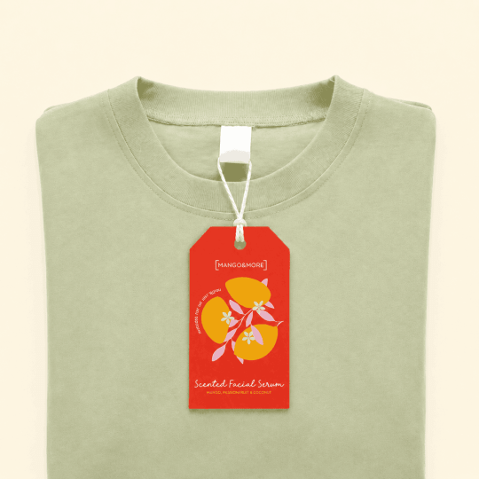 Złożona koszulka w kolorze szałwiowej zieleni leży na kremowym tle. Do kołnierzyka przyczepiona jest duża, czerwona metka z napisem '[MANGO & MORE]', 'Scented Facial Serum' oraz grafiką przedstawiającą owoce mango i białe kwiaty.