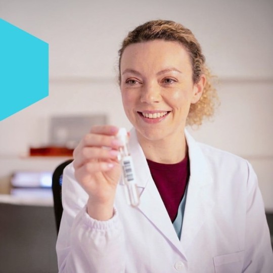 Uśmiechnięta pracowniczka laboratorium w białym kitlu, trzymająca w dłoni probówkę z naklejoną etykietą z kodem kreskowym.