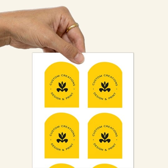 Dłoń trzymająca arkusz z naklejkami w kształcie łuku. Naklejki są żółte z czarnym logo i napisem 'CUSTOM CREATIONS DESIGN & PRINT'.