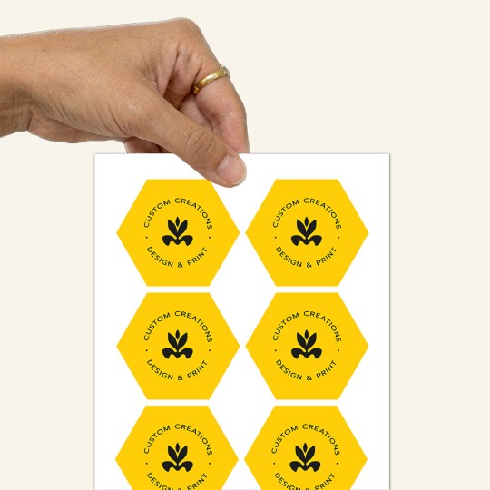 Dłoń trzymająca arkusz z naklejkami w kształcie sześciokąta. Naklejki są żółte z czarnym logo i napisem 'CUSTOM CREATIONS DESIGN & PRINT'.