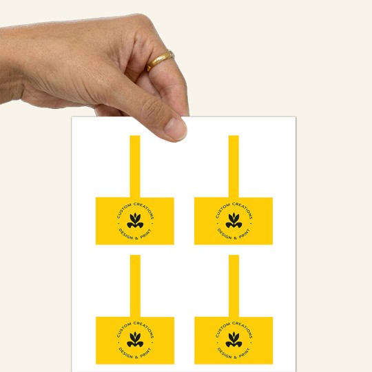 Ręka trzymająca arkusz z czterema żółtymi etykietami produktowymi. Każda etykieta składa się z kwadratowej części z czarnym logo i napisem "Custom Creations, Design & Print" oraz długiego paska do zaklejenia opakowania.