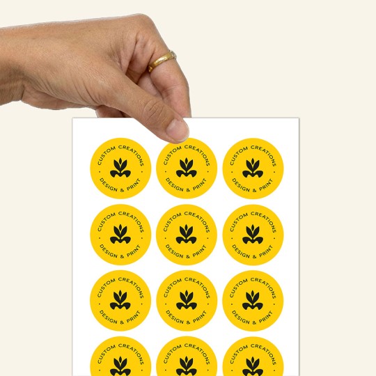 Dłoń trzymająca arkusz z okrągłymi naklejkami. Naklejki są żółte z czarnym logo i napisem 'CUSTOM CREATIONS DESIGN & PRINT'.