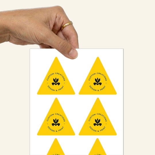 Dłoń trzymająca arkusz z naklejkami w kształcie trójkąta. Naklejki są żółte z czarnym logo i napisem 'CUSTOM CREATIONS DESIGN & PRINT'.