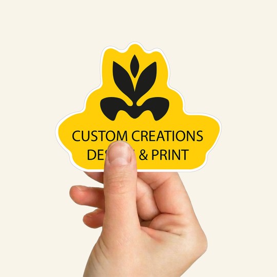 Dłoń trzymająca żółtą naklejkę o niestandardowym kształcie, z czarnym logo i napisem "Custom Creations Design & Print".