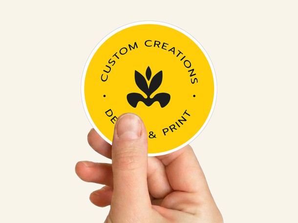 Dłoń trzymająca pojedynczą, okrągłą, żółtą naklejkę z czarnym logo i napisem "Custom Creations Design & Print".