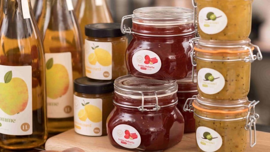Trzy butelki cydru z prostą etykietą z jabłkiem oraz dwa słoiki z dżemem truskawowym oraz z kiwi