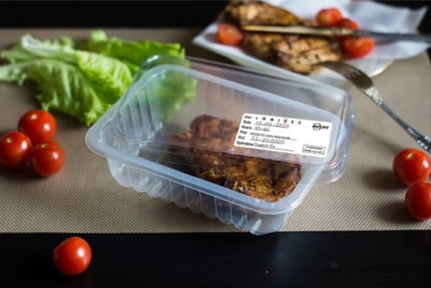 Plastikowy pojemnik z gotowym daniem i widoczną etykietą zawierającą datę przygotowania i termin ważności. W tle widoczne liście sałaty, pomidorki koktajlowe i talerz z jedzeniem.