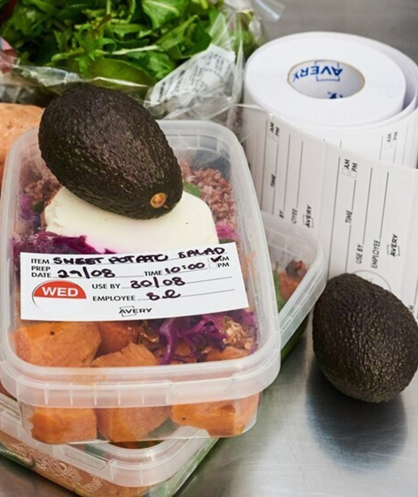 Pojemnik z sałatką z batatów, na którym umieszczona jest wyraźna etykieta z datą przygotowania, datą przydatności i informacjami o produkcie. Na pojemniku leży awokado, w tle rolka etykiet i inne produkty spożywcze.