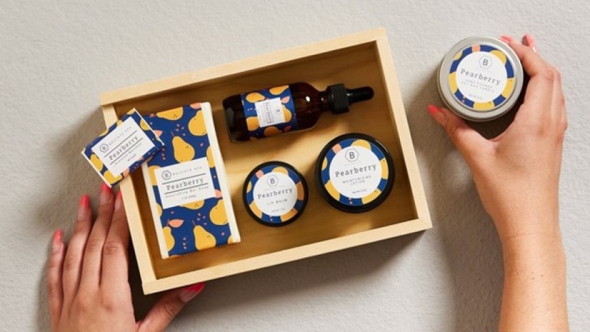 Zdjęcie przedstawia zestaw kosmetyków Pearberry o spójnym, kolorowym designie z motywem gruszek na granatowym tle. W drewnianym pudełku znajdują się: prostokątne mydło w papierze, szklana butelka z pipetą, dwie okrągłe puszki oraz mała kartonowa etykieta. Poza pudełkiem, po prawej stronie, osoba trzyma w dłoni kolejny okrągły kosmetyk z tej samej serii. Tło jest jasnoszare, dłonie mają pomalowane paznokcie na różowo.