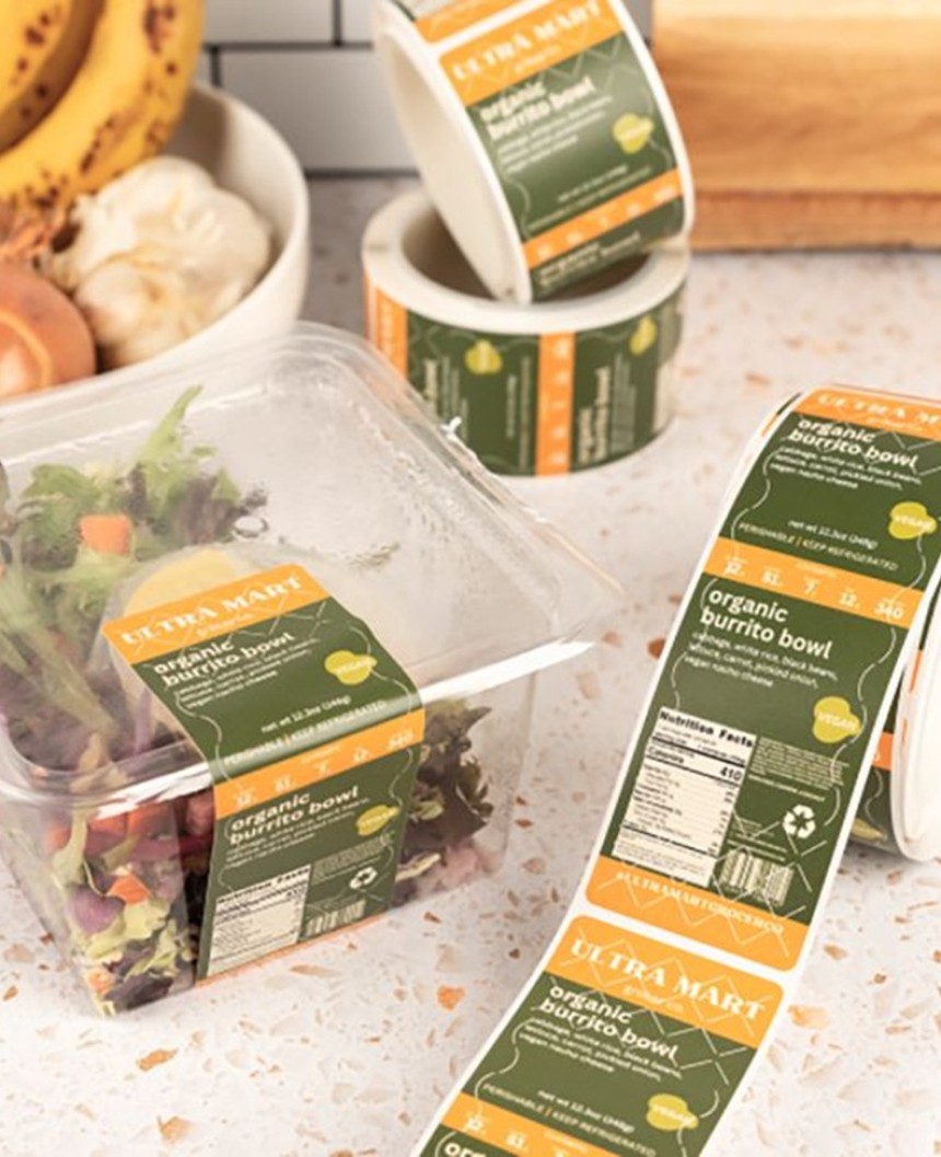 Przezroczyste pojemniki z sałatką z etykietą „organic burrito bowl” i rolki podobnych zielono-pomarańczowych nalepek, ustawione na jasnym blacie kuchennym.