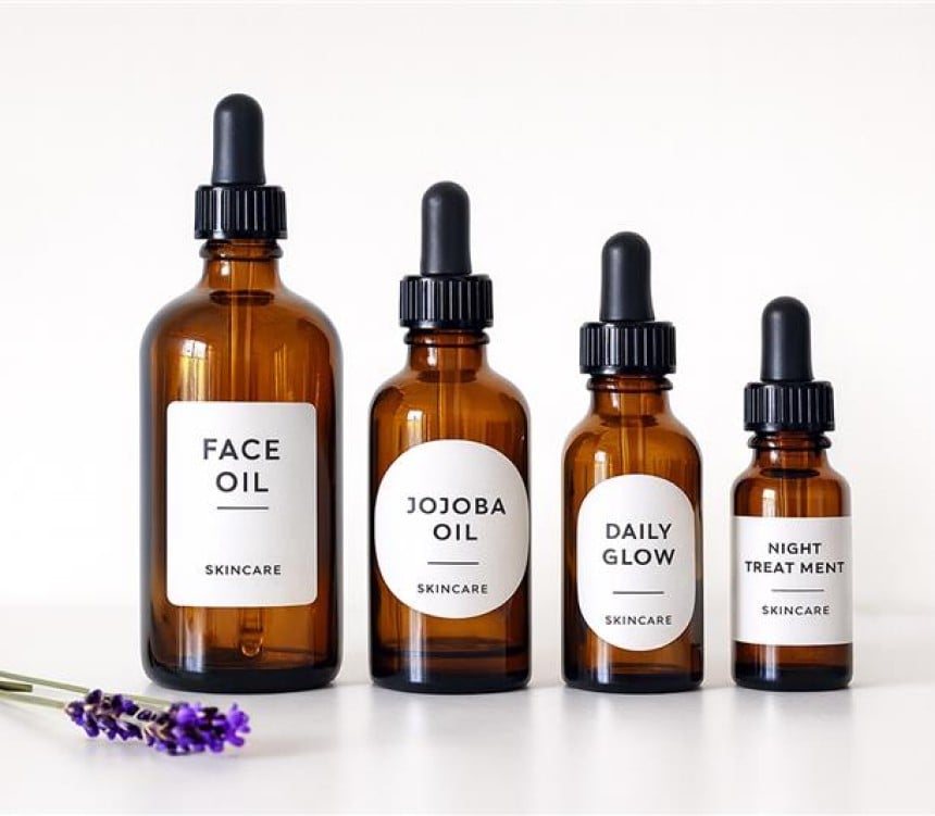Cztery brązowe butelki z pipetą do pielęgnacji twarzy ustawione w rzędzie na białym tle; etykiety opisane kolejno: „Face Oil”, „Jojoba Oil”, „Daily Glow” i „Night Treatment”, z drobnym dopiskiem „Skincare”; z lewej leży gałązka lawendy.