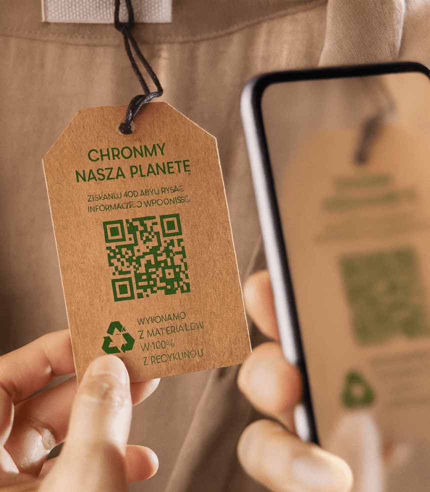 Zbliżenie na metkę z brązowego kartonu przymocowaną do ubrania. Na metce zielony napis „CHROŃMY NASZĄ PLANETĘ”, niżej kod QR i informacja, że produkt wykonano w 100% z materiałów z recyklingu. Po prawej fragment telefonu, którym ktoś skanuje kod.