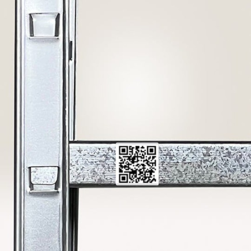Fragment metalowego regału magazynowego z małą, białą etykietą z kodem QR naklejoną na poziomą belkę, przeznaczoną do skanowania lokalizacji lub produktu.